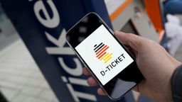 Eine Hand hält ein Smartphone auf dem ein Deutschland-Piktogramm mit der Unterschrift "D-TICKET" zu sehen ist vor einen Fahrscheinautomaten | Bild:picture alliance / Flashpic | Jens Krick