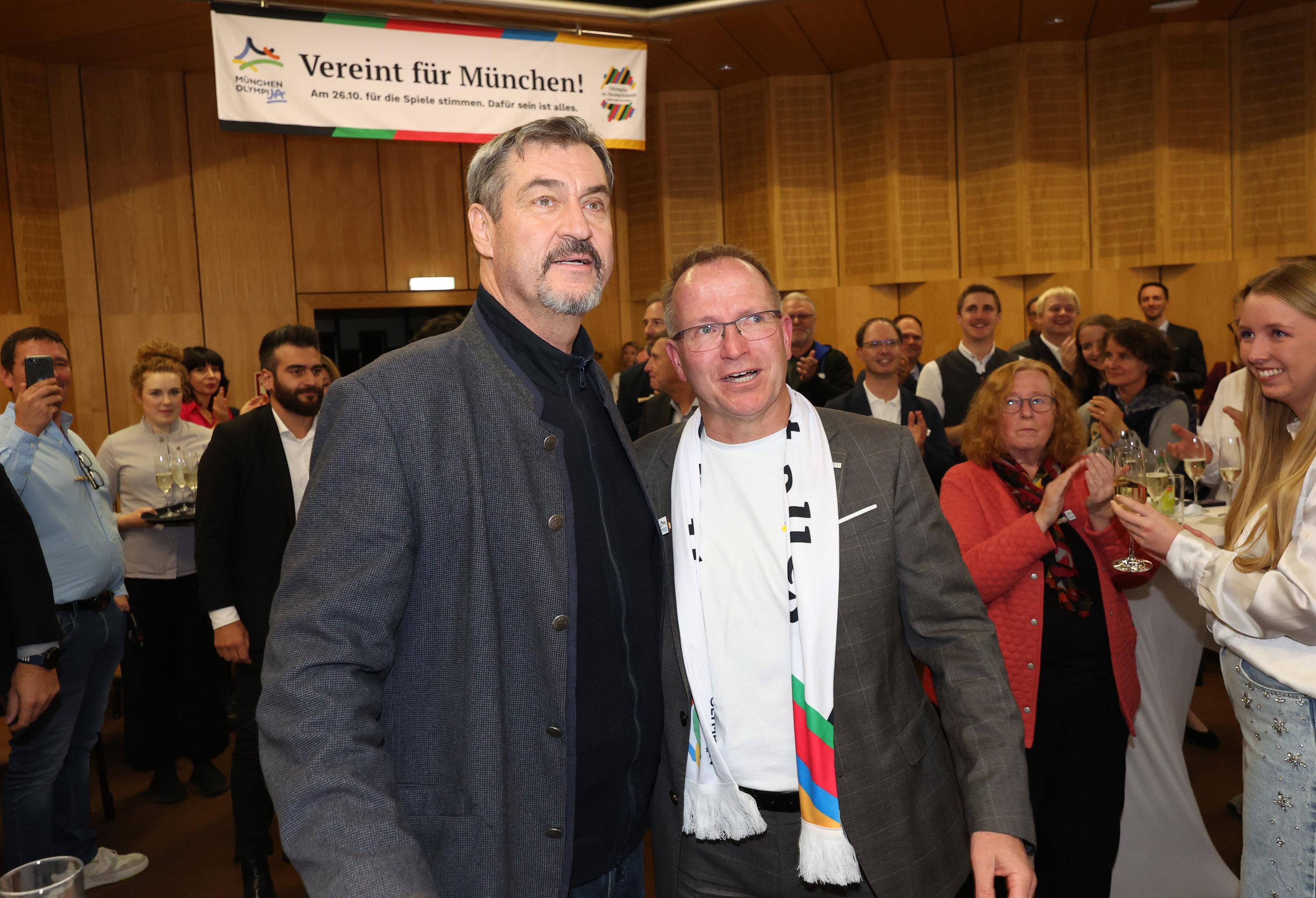 26.10.2025, Bayern, München: Markus Söder (CSU, l), Ministerpräsident von Bayern, und Jörg Ammon, Präsident des Bayerischen Landes-Sportverbands (BLSV), verfolgen die Bekanntgabe der ersten Hochrechnungen zum Ergebnis des Bürgerentscheids zur Olympia-Bewerbung auf einer Leinwand. Bei der Bürgerbefragung stimmten nach Angaben von Münchens Oberbürgermeister Reiter deutlich über 60 Prozent für eine Bewerbung der bayerischen Landeshauptstadt um Sommerspiele und Paralympische Spiele im Jahr 2036, 2040 oder 2044. 