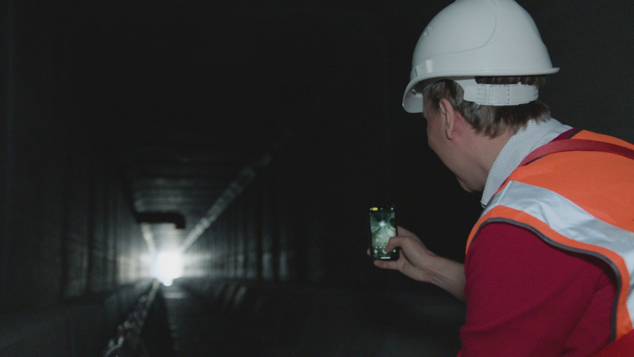 Ein Mann fotografiert mit einem Smartphone in der Hand in einen dunklen Tunnel, an dessen Ende es hell ist.