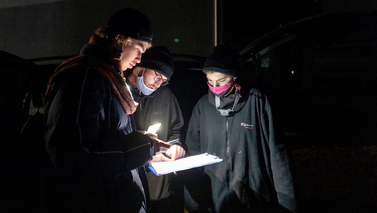 Im dunkeln stehen drei Menschen um ein Klemmbrett herum, dass sie mit einer Stirnlampe  beleuchten. 