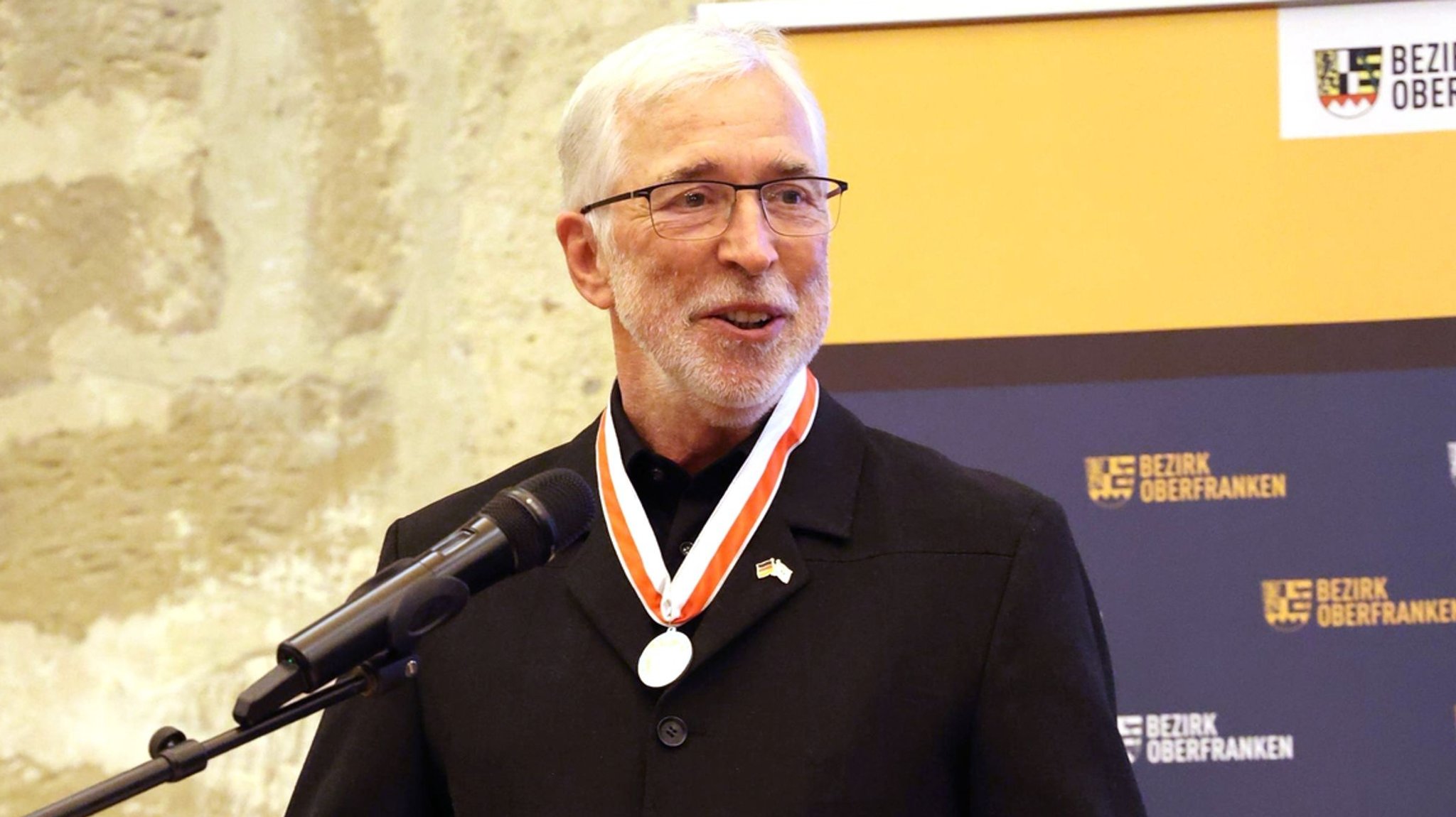 Michael Genniges auf dem Podium. Um den Hals trägt er die Silberne Ehrenmedaille des Bezirks Oberfranken, der er jetzt zurückgegeben hat