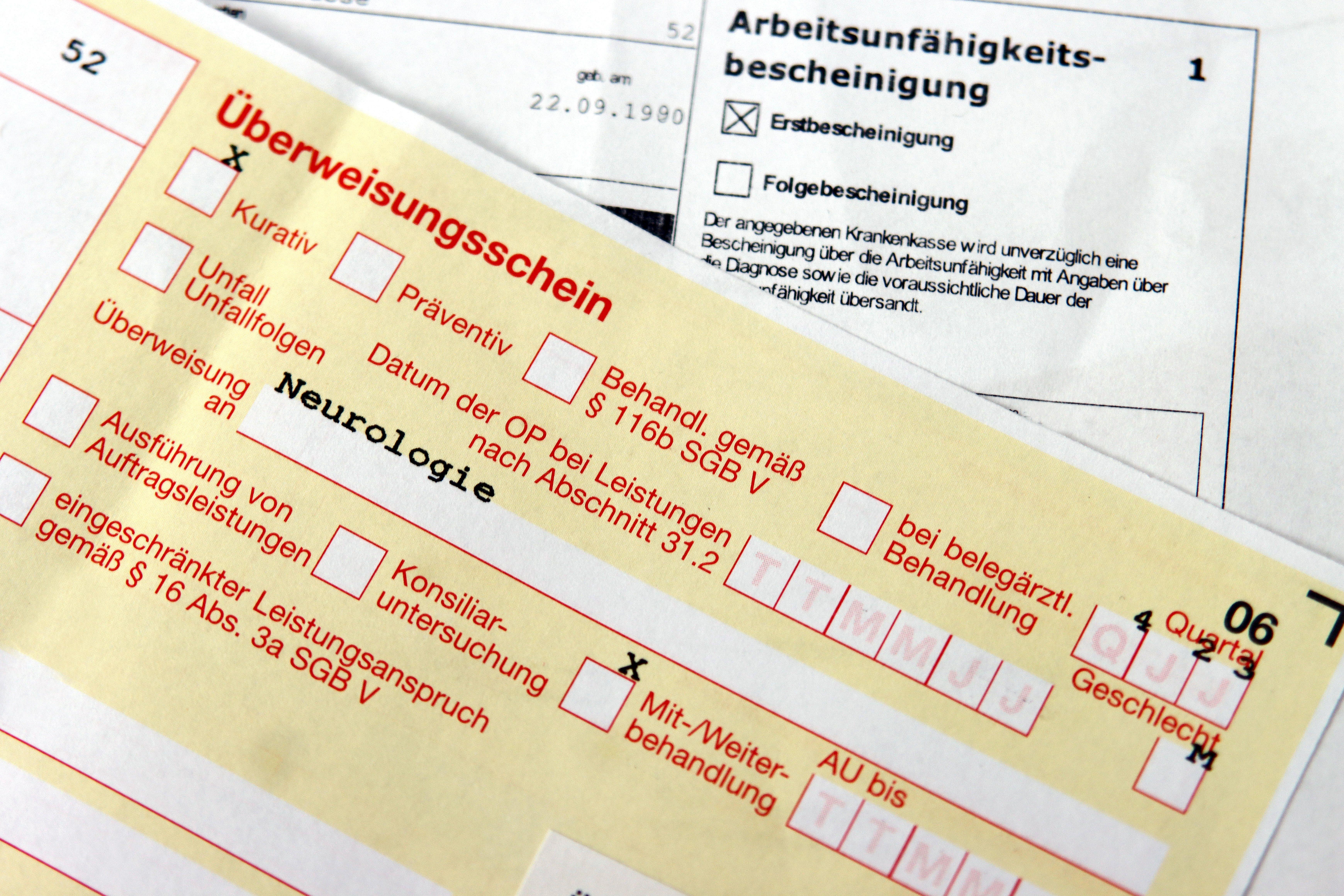 (Symbolbild) Ein gelber Überweisungsschein zur Neurologie, im Hintergrund eine Arbeitsunfähigkeitsbescheinigung