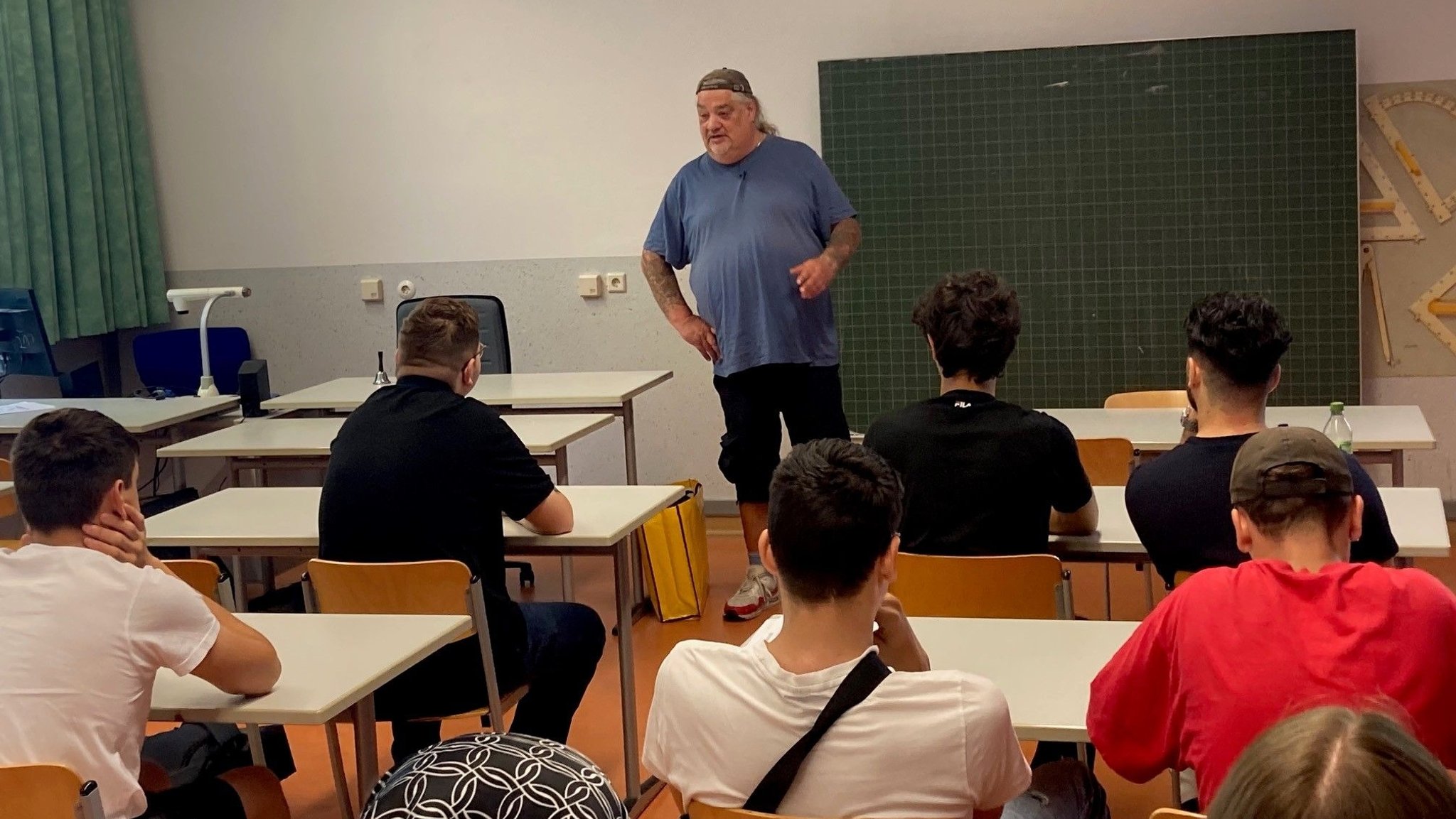 Ein älterer Mann mit langen grauen Haaren, einem blauen Shirt und einer Cap steht in einem Klassenzimmer vor einer Schulklasse.