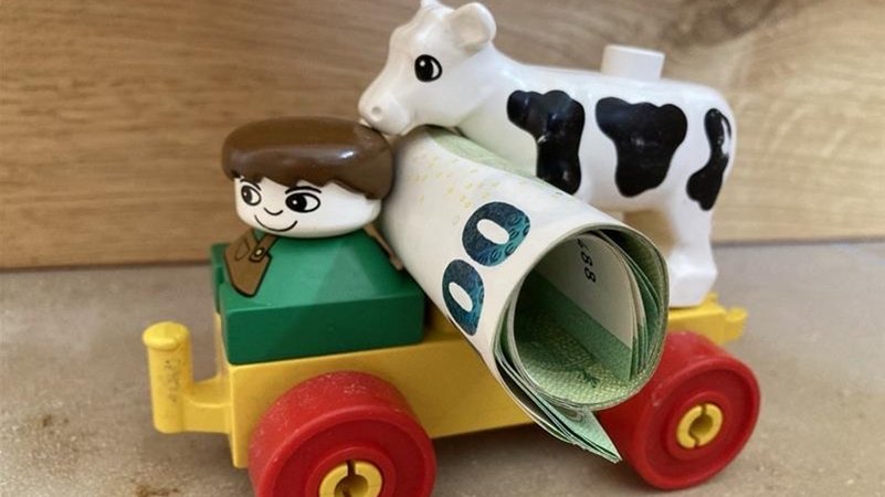 Spielzeuglandwirt, der auf einem Wagen sitzend Geld und eine Kuh transportiert