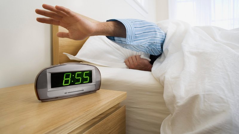 Ein Arm wird aus dem Bett gestreckt und ist kurz davor, auf einen Digitalwecker zu schlagen. | Bild: picture alliance / Design Pics / First Light | Bruno Crescia Ein Arm wird aus dem Bett gestreckt und ist kurz davor, auf einen Digitalwecker zu schlagen.