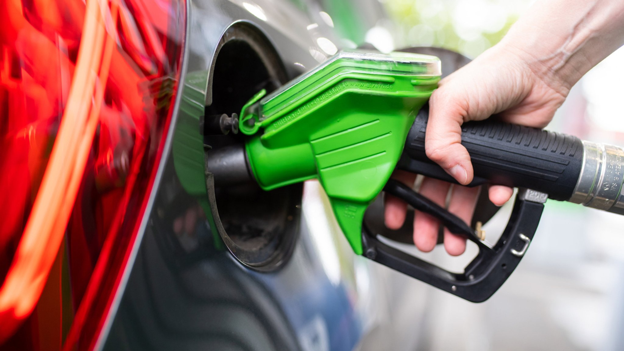 Eine Frau hält an einer Tankstelle an einer Zapfsäule eine Zapfpistole in der Hand und betankt ein Auto.