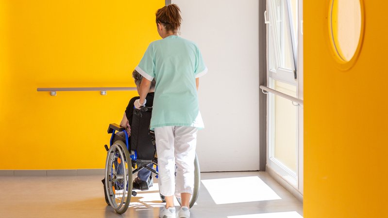 Eine Altenpflegerin schiebt eine im Rollstuhl sitzende Person (Symbolbild) | Bild: stock.adobe.com/Reddragonfly Eine Altenpflegerin schiebt eine im Rollstuhl sitzende Person (Symbolbild)