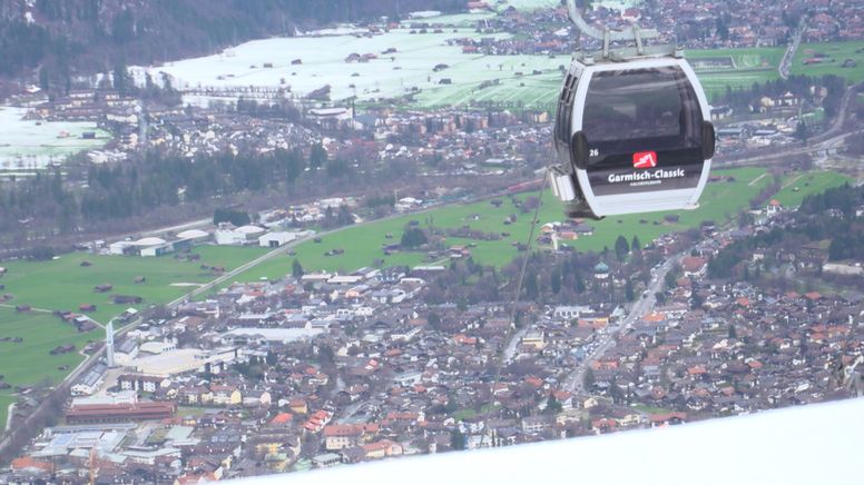 Im Hintergrund grüne Wiesen und Garmisch-Partenkirchen - im Vordergrund Schnee und eine Gondel. | Bild:BR/Martin Breitkopf