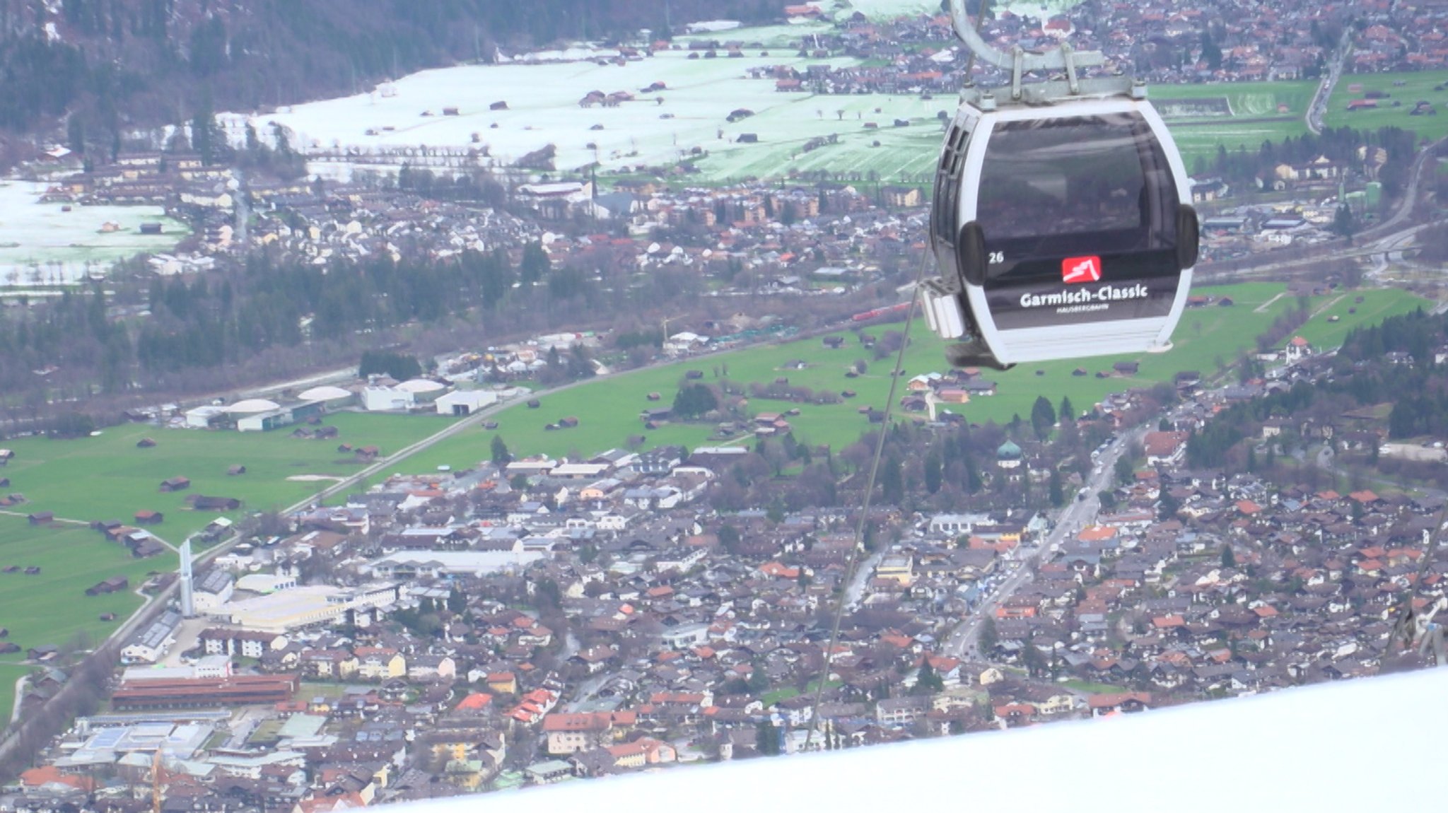 Im Hintergrund grüne Wiesen und Garmisch-Partenkirchen - im Vordergrund Schnee und eine Gondel.