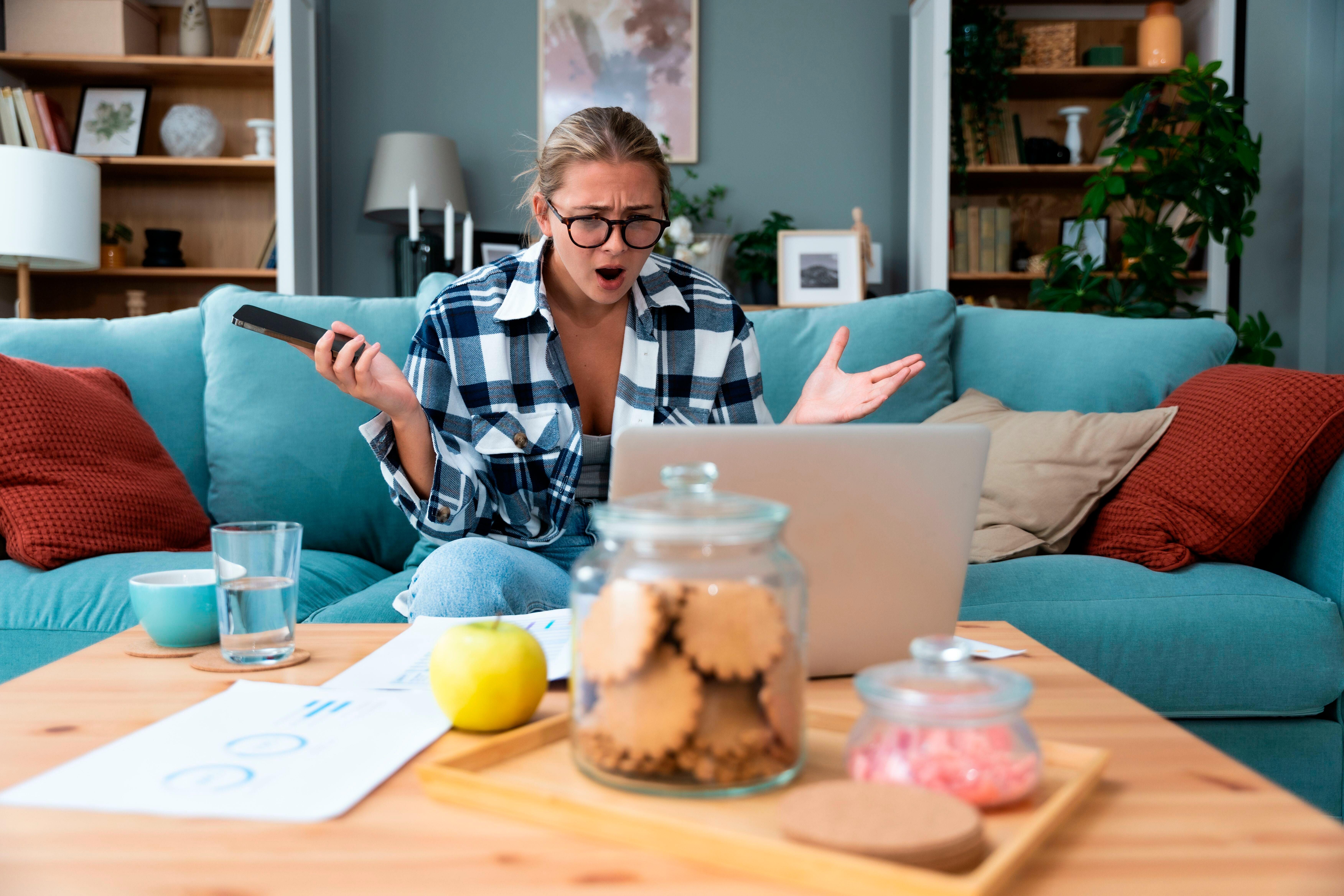 Symbolbild: Eine Frau sitzt auf einer Couch vor einem Laptop, mit dem Smartphone in der Hand und chaotisch arrangierten Gegenständen auf dem Tisch. Sie wirkt abgelenkt und wütend.