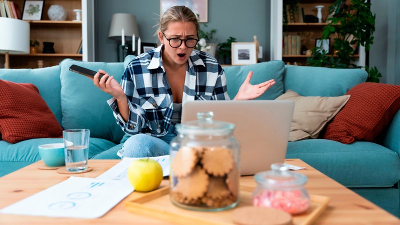 Symbolbild: Eine Frau sitzt auf einer Couch vor einem Laptop, mit dem Smartphone in der Hand und chaotisch arrangierten Gegenständen auf dem Tisch. Sie wirkt abgelenkt und wütend. | Bild: picture alliance / Visually | Srdjan Randjelovic Symbolbild: Eine Frau sitzt auf einer Couch vor einem Laptop, mit dem Smartphone in der Hand und chaotisch arrangierten Gegenständen auf dem Tisch. Sie wirkt abgelenkt und wütend.