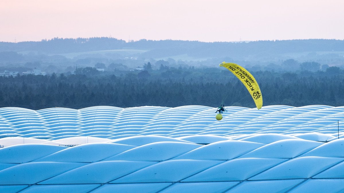 Der Angeklagte in seinem gelben Segelflieger mit einem großen gelben Ball. Er segelt über dem Dach der Allianz Arena mit seinen rautenförmigen Luftkissen, die im Abendlicht bläulich leuchten.