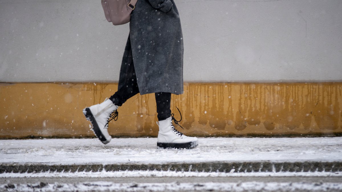 Symbolbild: Eine Person mit weißen Stiefeln, Mantel und Rucksack läuft auf einem von Schnee bedeckten Gehweg.