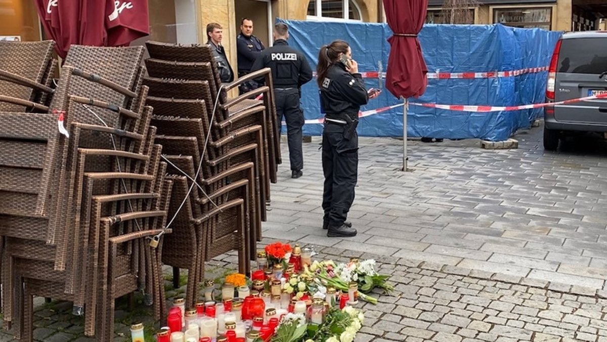 Polizeibeamte in Uniform stehen in einem Innenhof und telefonieren, am Boden sind Blumen und Kerzen. 