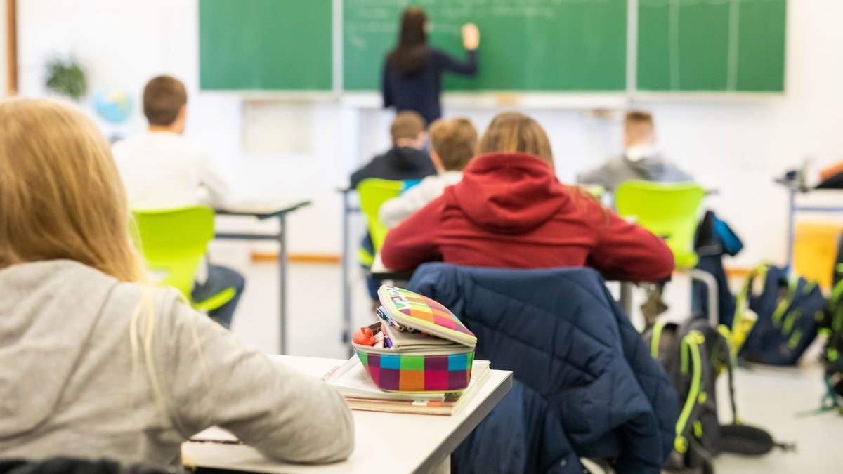 Blick in ein Klassenzimmer: Mehrere Kinder sitzen an ihren Plätzen während eine Lehrerin etwas auf die Tafel schreibt.