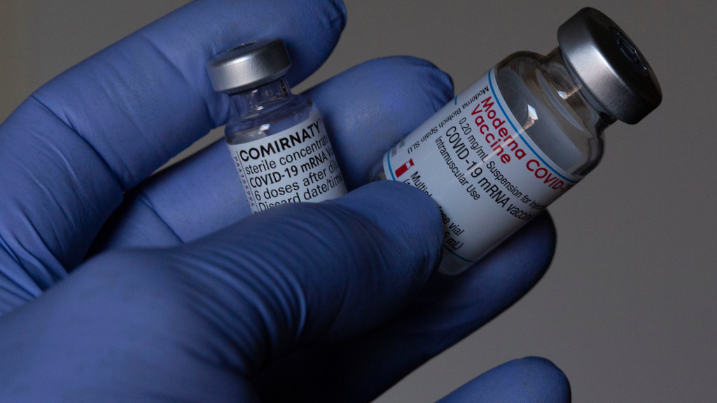 Hand in in blauem Kunststoffhandschuh hält je eine Ampulle der Impfstoffe von Biontech und Moderna.