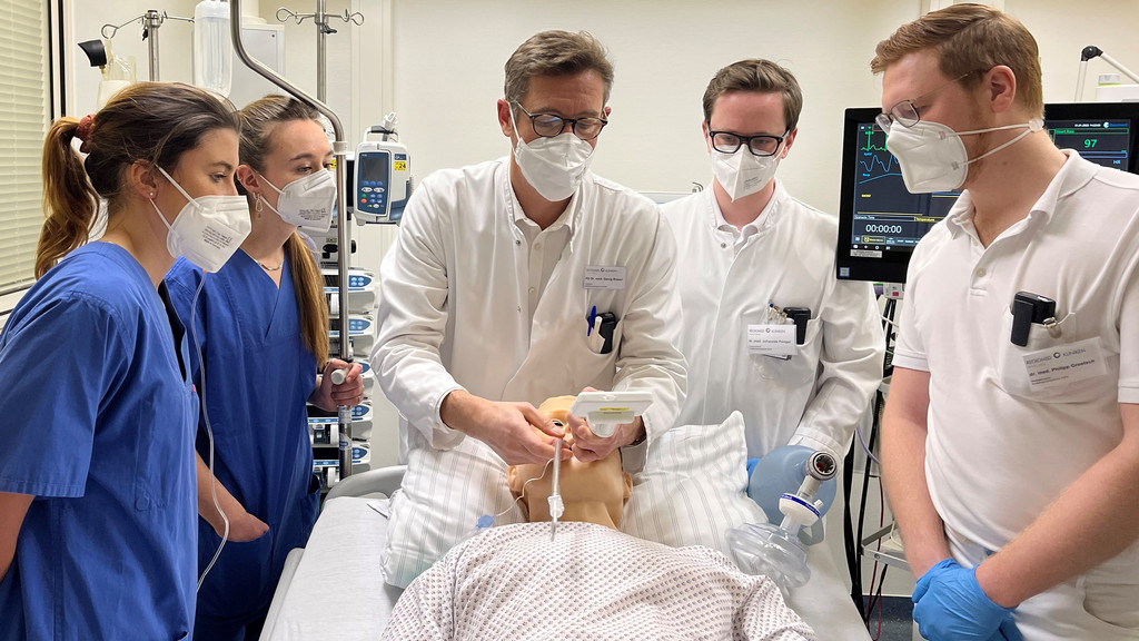 Ein Arzt schiebt einer Simulator-Puppe einen Beatmungsschlauch in den Hals, vier Medizinstudenten stehen am Krankenbett und schauen zu. 