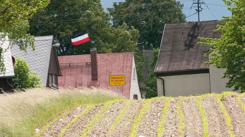 Das gelbe Ortsschild mit der Aufschrift "Oberprex", dahinter Häuser mit einer schwarz-weiß-roten Fahne, im Vordergrund ein Feld. 