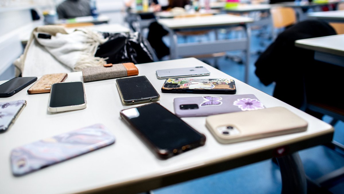 Handys liegen im Vordergrund auf einem Tisch im Klassenzimmer.