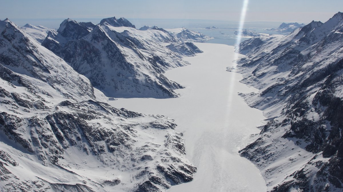 Fjord in Südost-Grönland, schneebedeckt und eingefroren, am Horizont ist das Meer. 