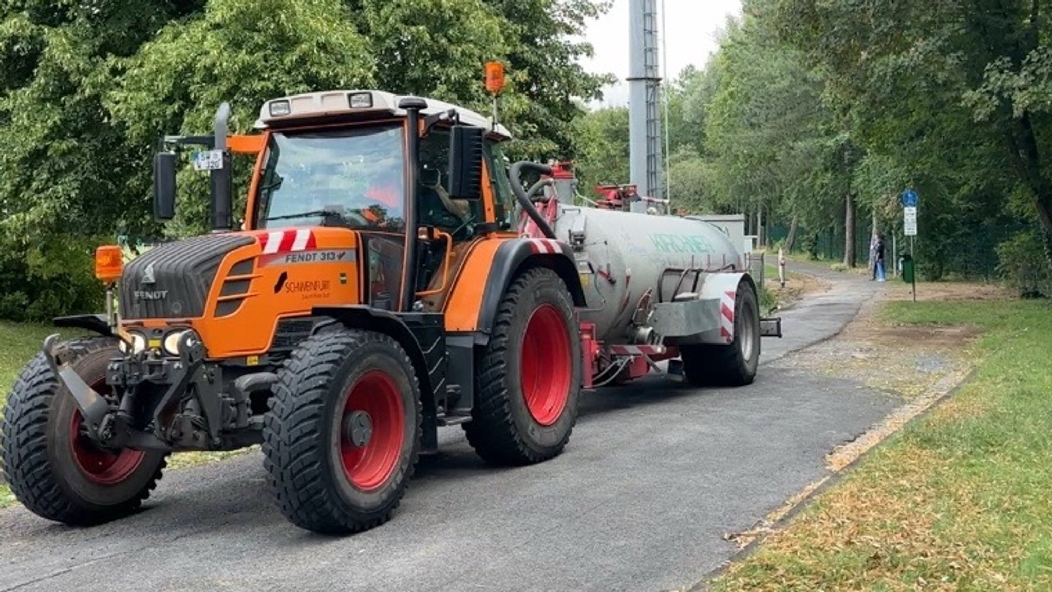 Ein Traktor mit einem Tankanhänger steht an einem Weg. 