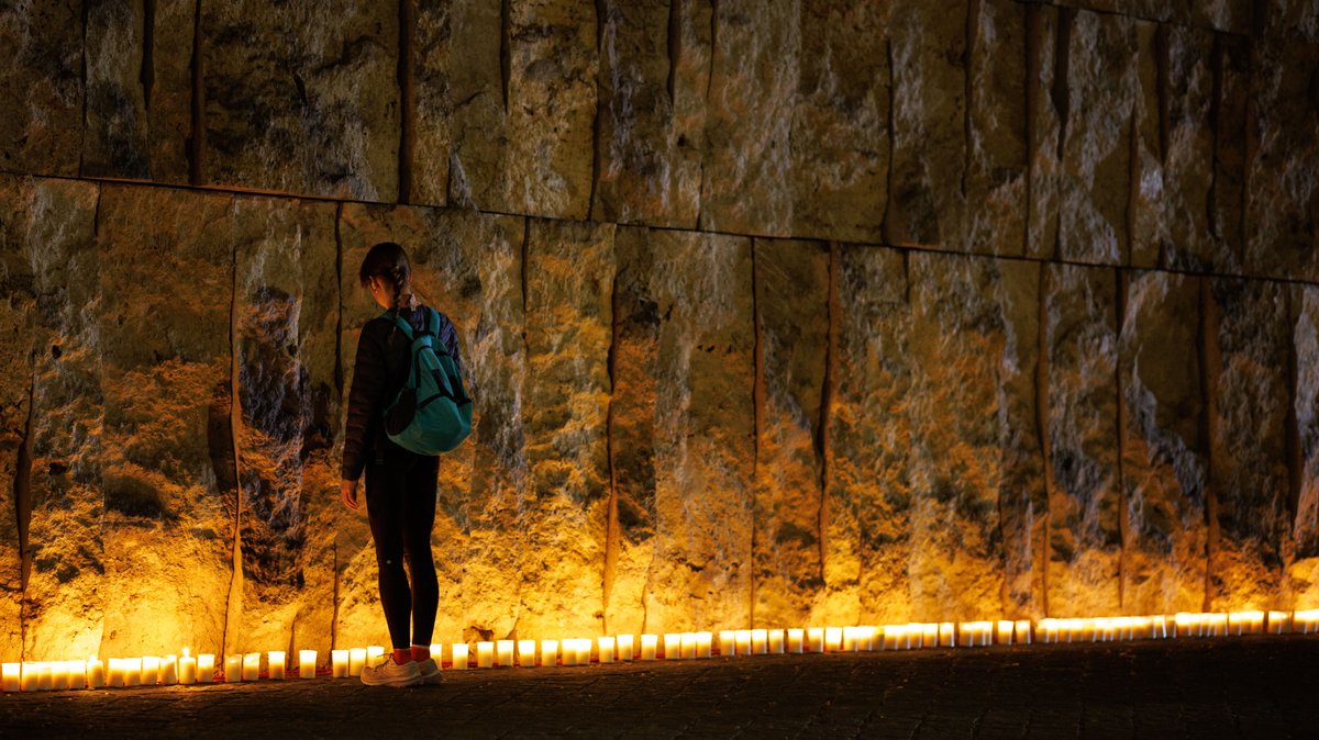 Eine junge Frau steht bei einer Gedenkveranstaltung der Israelitischen Kultusgemeinde München und Oberbayern vor der Münchner Hauptsynagoge Ohel Jakob an der Außenfassade, vor der hunderte Kerzen angezündet wurden.