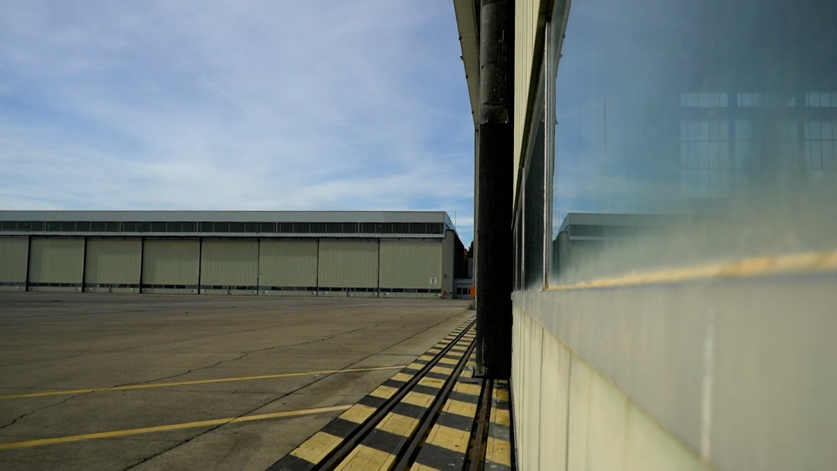Blick auf das Gelände des ehemaligen Militärflughafens: ein riesiger leerer Platz, im Anschnitt und im Hintergrund zwei große Hangars.