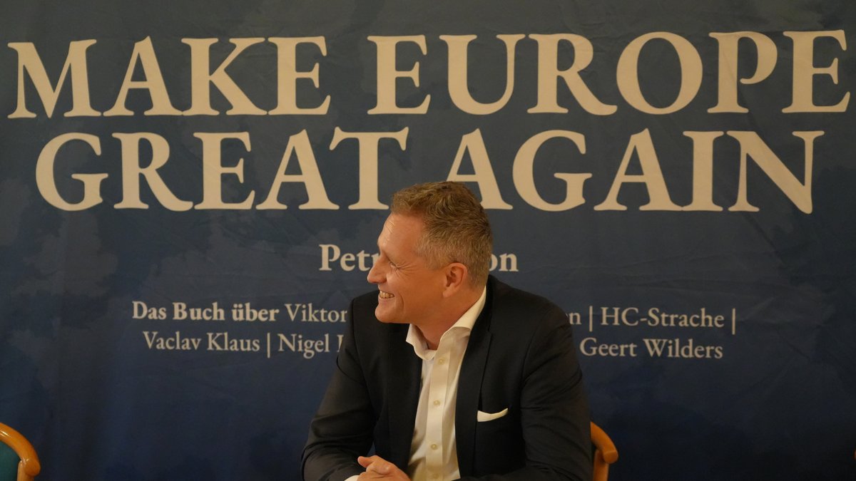 Petr Bysrton sitzt vor einem großen Banner, auf dem steht: "Make Europe Great Again"