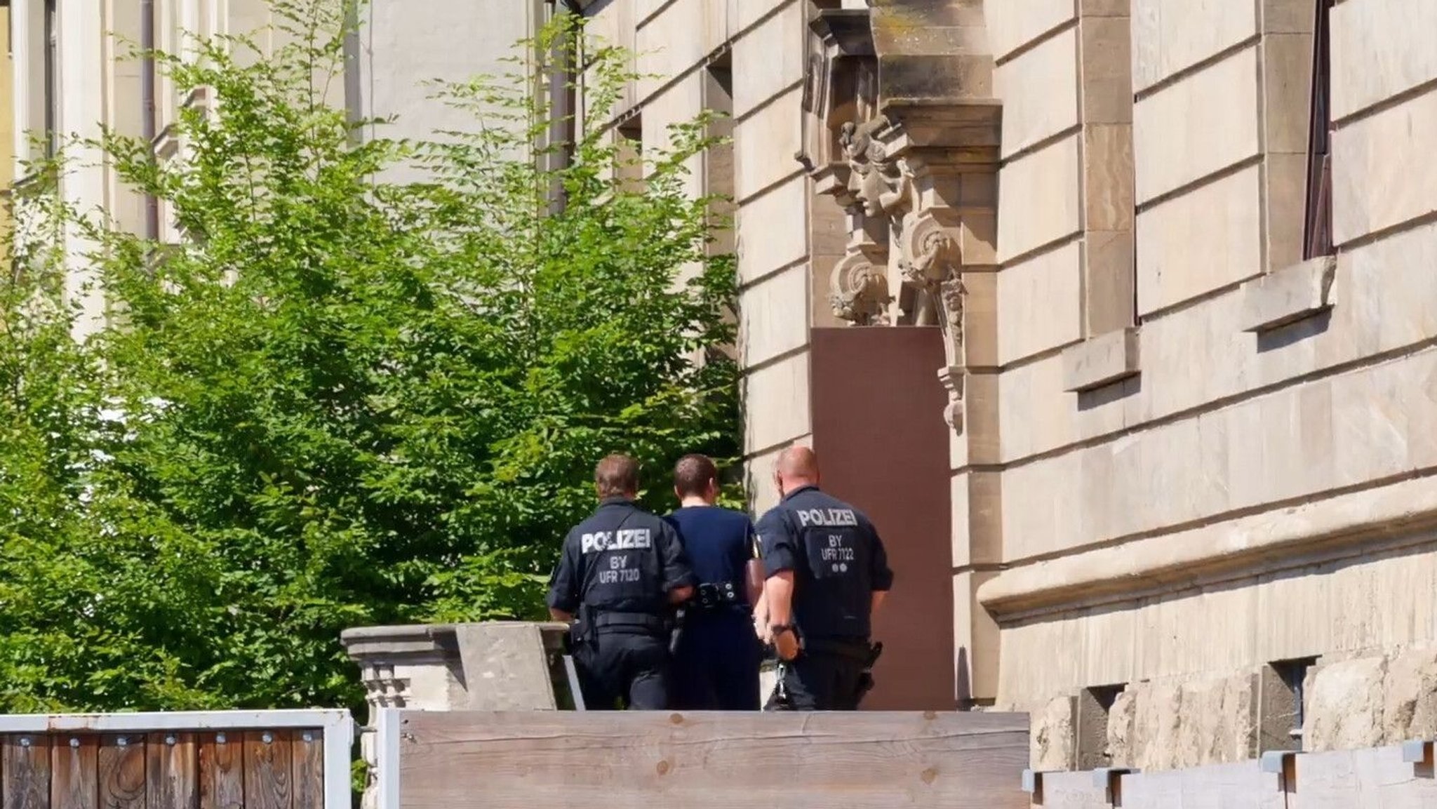 Archivbild 2. Juli 2025: Das Standbild aus einem Video zeigt, wie Polizeibeamte den Verdächtigen, der am Dienstag beim Stromversorger Überlandwerk Rhön in Mellrichstadt Kollegen mit einem Messer angegriffen, verletzt und eine tödlich verletzt haben soll, ins Gerichtsgebäude bringen. 