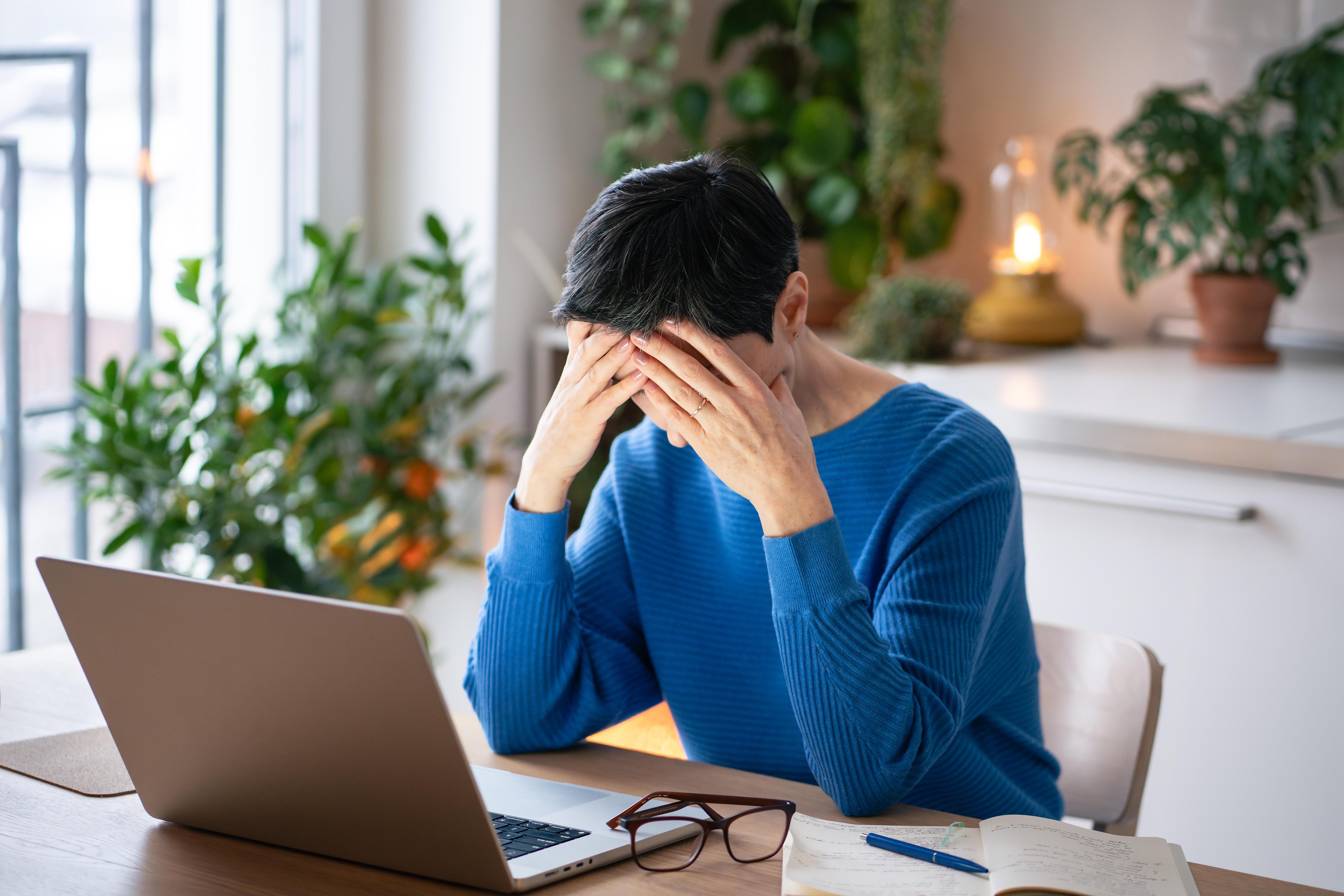 Eine Frau im blauen Pullover sitzt vor einem Laptop und stützt den Kopf in die Hände
