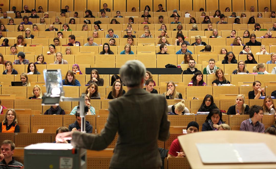 (Symbolbild) Eine Professorin steht vor einem fast vollbesetzten Hörsaal, in dem die meisten Studierenden zu ihr blicken.