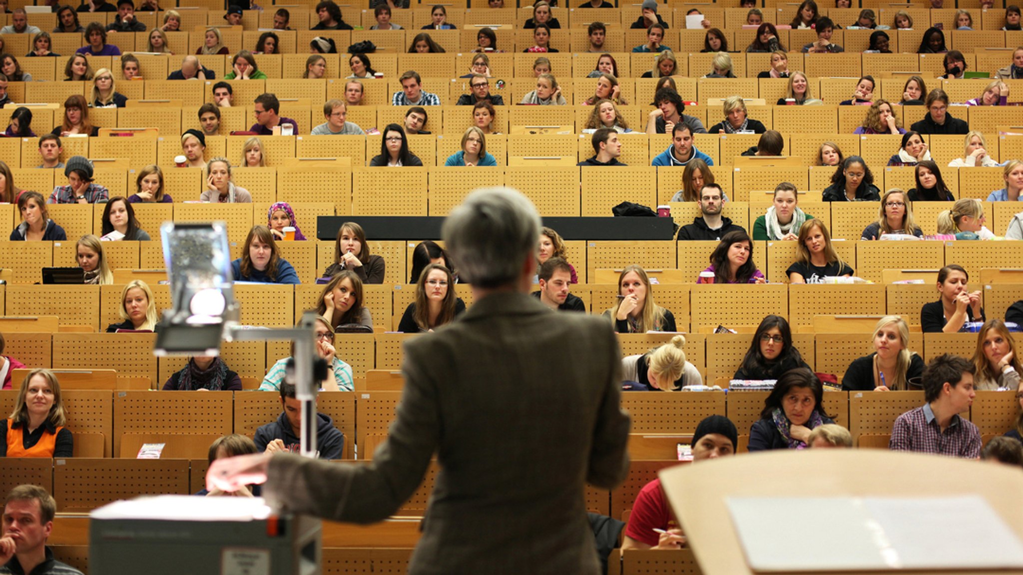 (Symbolbild) Eine Professorin steht vor einem fast vollbesetzten Hörsaal, in dem die meisten Studierenden zu ihr blicken.