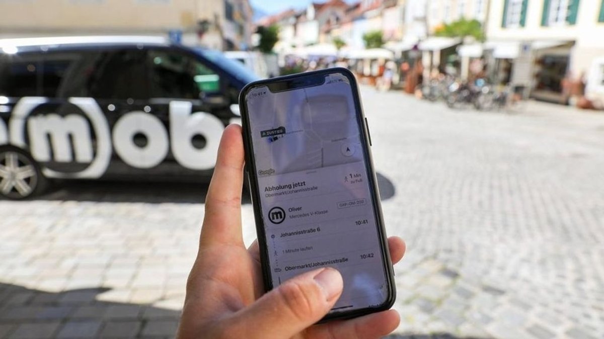 Omobi-Rufbus in Murnau, davor ein Handy, auf dessen Bildschirm eine Rufbus-Bestellung angezeigt wird.