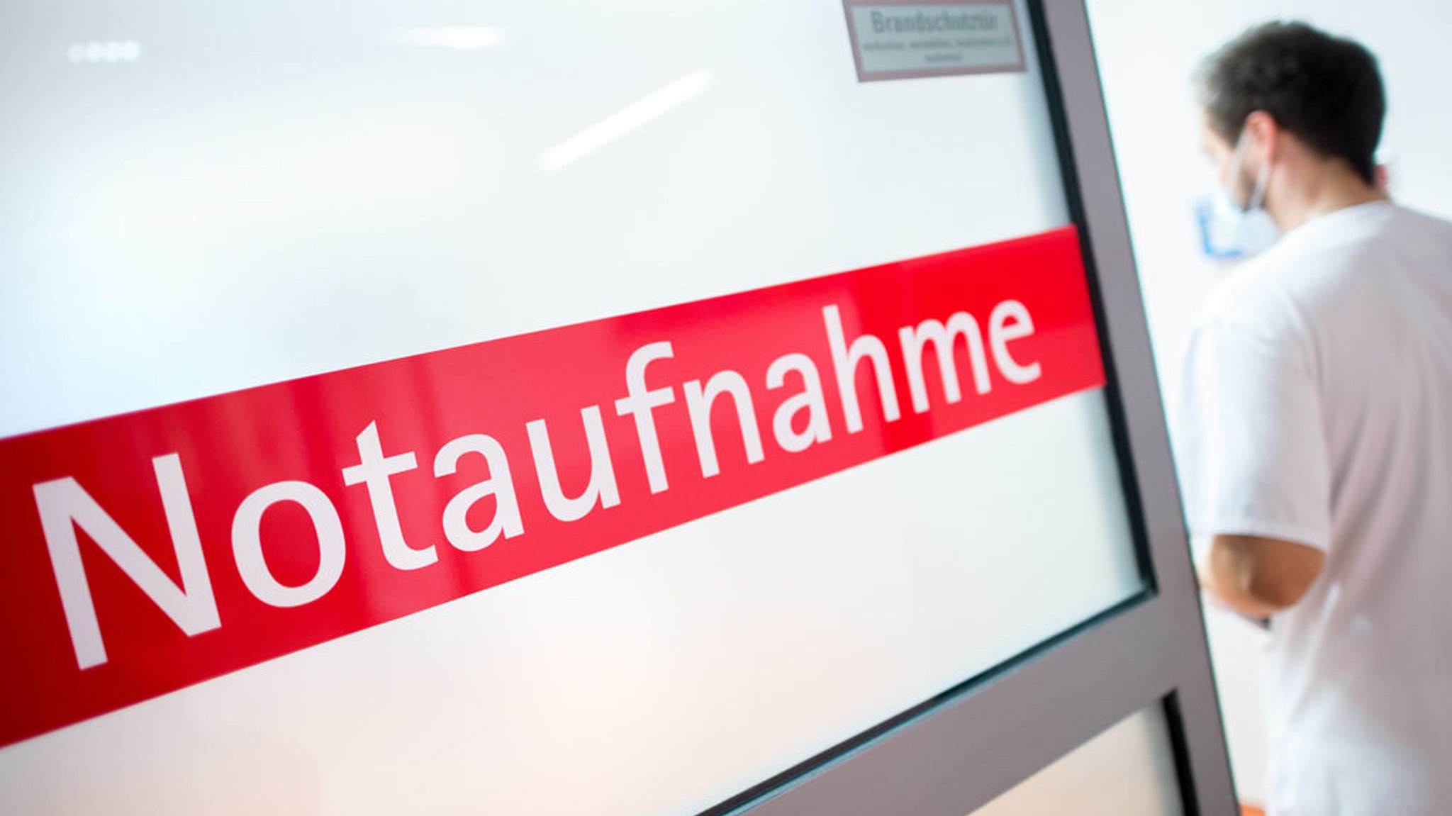 Ein Krankenpfleger steht in einem Krankenhaus neben einer Glastür mit der Aufschrift "Notaufnahme". 