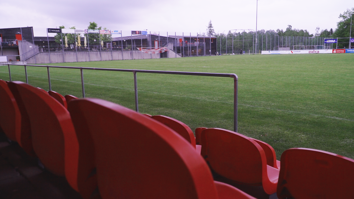 Man sieht die Sitze einer Zuschauertribüne und im Hintergrund den Fußballplatz