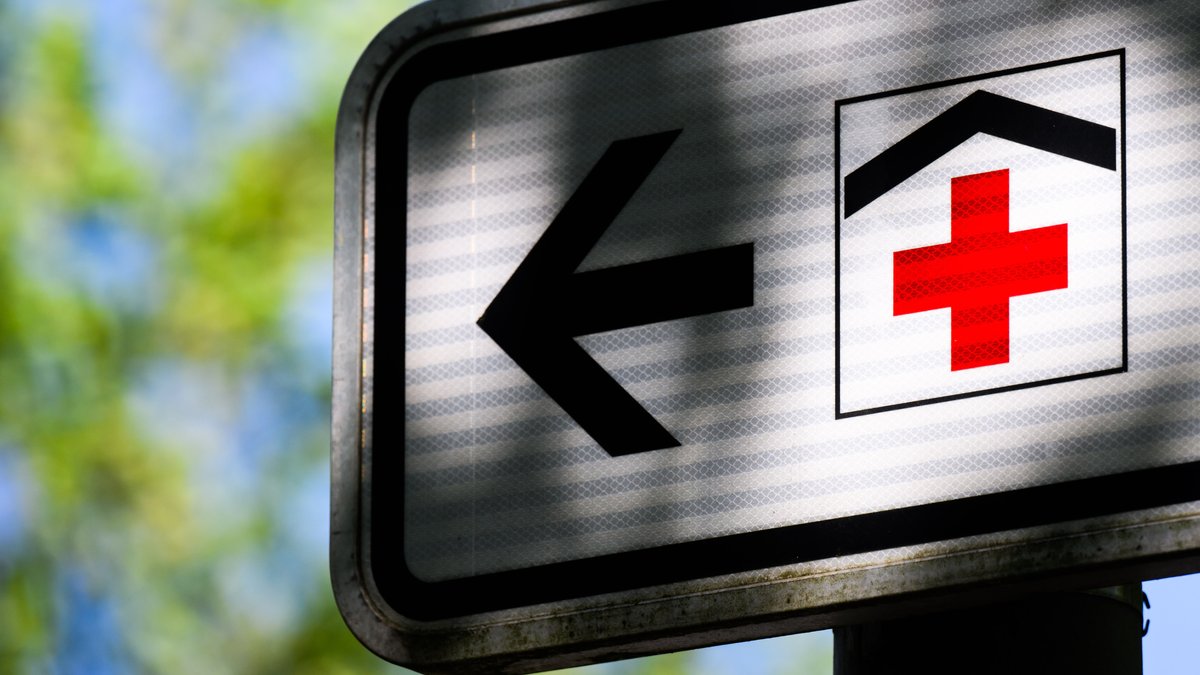 Straßenschild Krankenhaus mit einem roten Kreuz auf weißen Hintergrund mit einem schwarzen Dach darüber, daneben ein schwarzer Pfeil, der nach rechts zeigt.