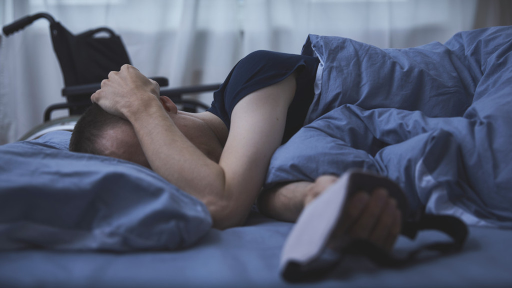 Mann liegt mit Schlafmaske in der Hand im Bett in einem abgedunkelten Zimmer, im Hintergrund steht ein Rollstuhl