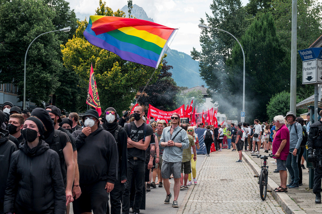 26.06.2022, Bayern, Garmisch-Partenkirchen: Schwarz gekleidete Demonstranten schwenken während der Demonstration gegen den G7 Gipfel die Gay Pride Flagge. 