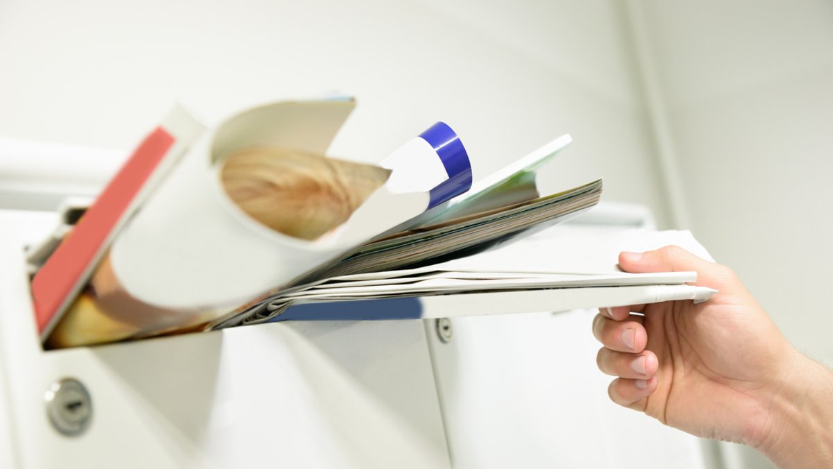 Eine Hand hält Post aus einem überfüllten Briefkasten.
