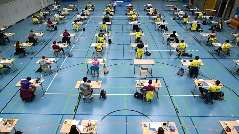 Schülerinnen und Schüler sitzen in einer Turnhalle an Tischen und schreiben ihre Abiturprüfung.