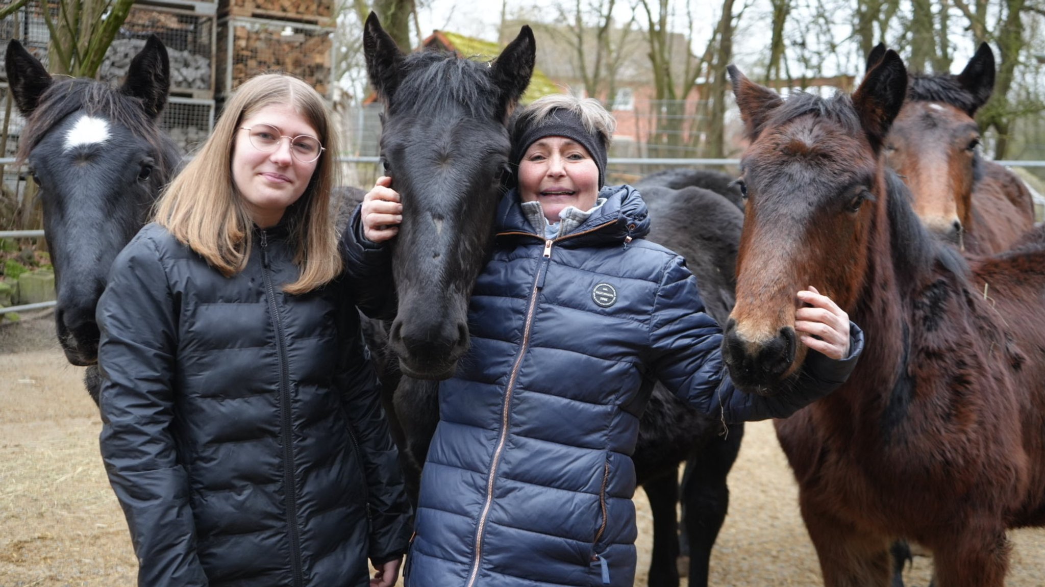 Zwei Frauen lachen umgeben von Pferden in die Kamera.