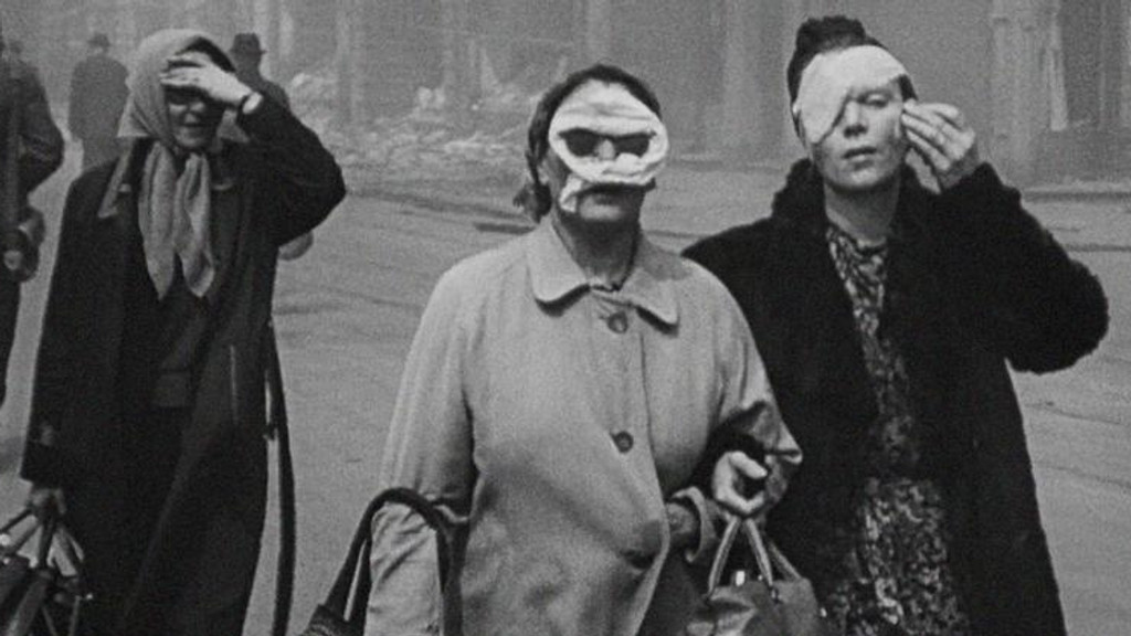Zwei Frauen mit Gesichts-Verbänden gehen auf historischem Schwarz-Weiß-Foto durch eine bombardierte Straße, hinter ihnen sind weitere Passanten zu sehen.     