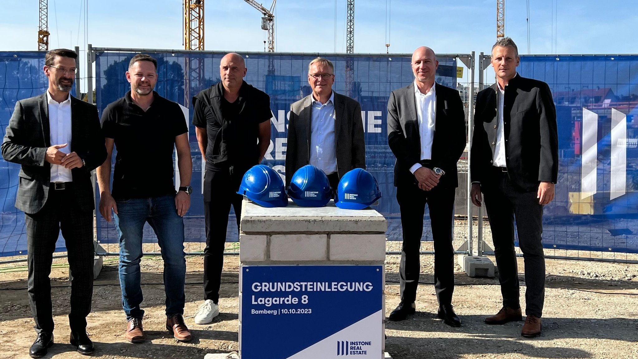 Sechs Männer stehen an einem Bauzaun auf einer Baustelle vor einer quadratischen, kleinen Mauer. Auf dieser liegen drei blaue Helmen. Auf der Mauer steht "Grundsteinlegung Lagarde 8"