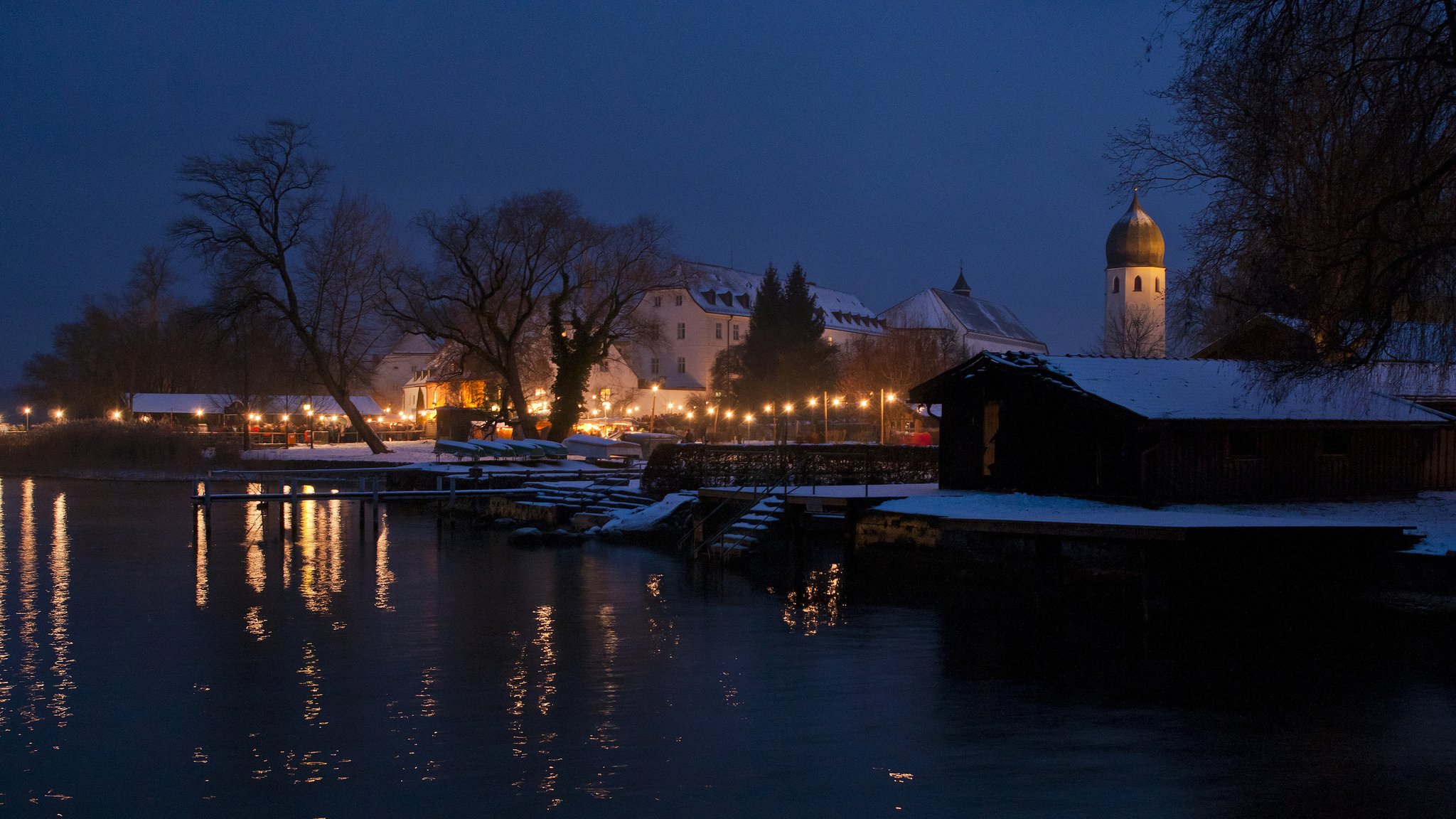 Blick auf die verschneite Insel Frauenchiemsee im Dunklen; Kirche und ein Weg sind mit Lichterketten beleuchtet. Der Christkindlmarkt auf Frauenchiemsee, einer der bekanntesten Weihnachtsmärkte Bayerns, stand vor dem Aus - doch jetzt ist er gerettet. (Archivbild)