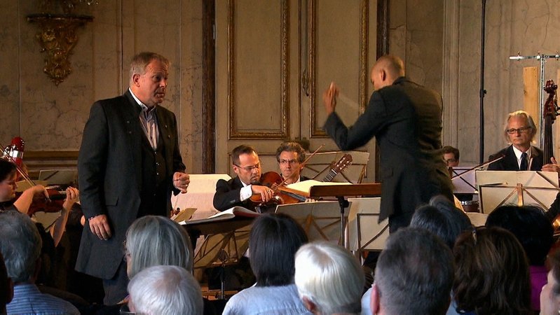 Ein Sänger im schwarzen Anzug bei einem Konzert, ein Dirigent leitet ein Orchester an.