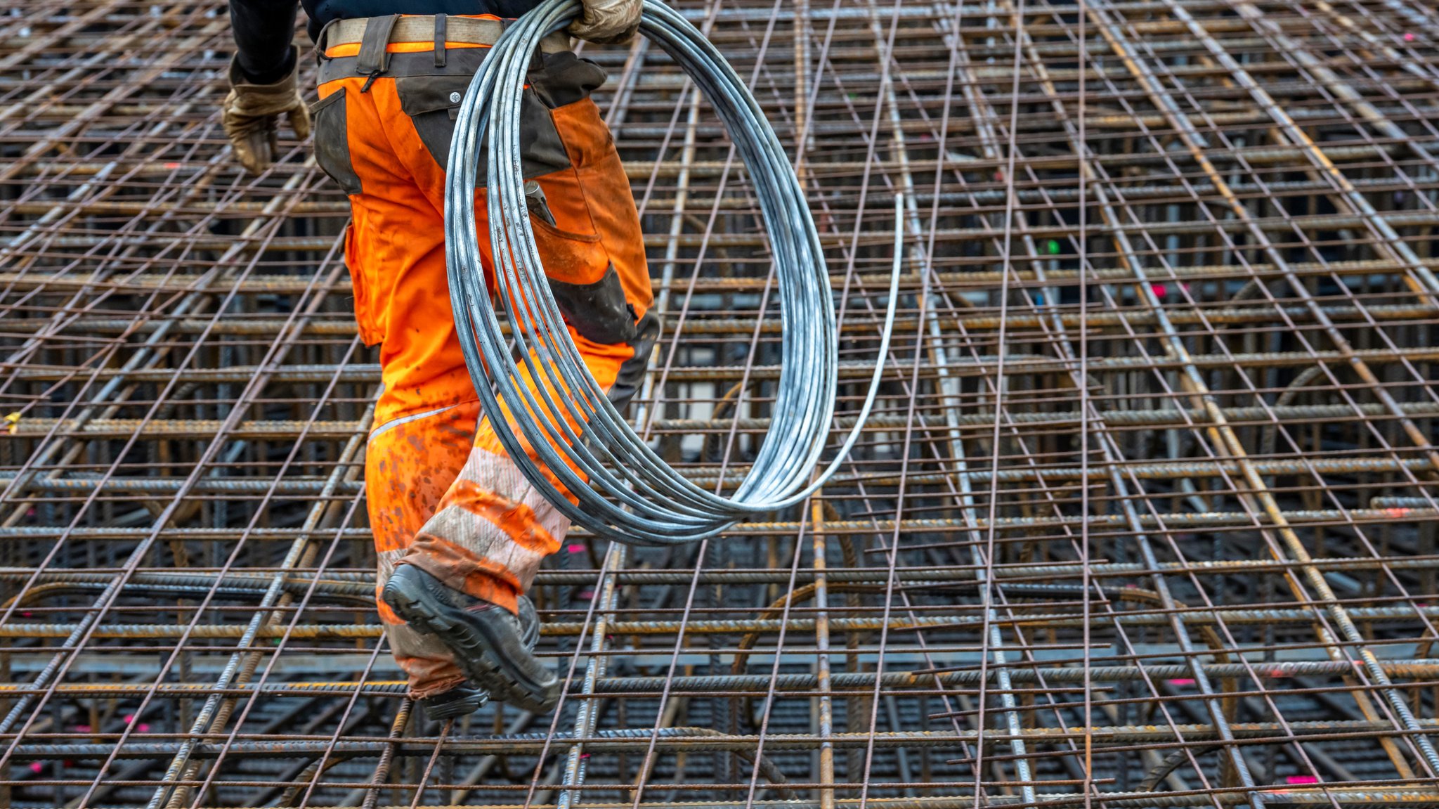 Ein Mann in orangenen Sicherheitsklamotten mit Reflektoren läuft auf einer Baustelle auf einem Gitter herum. Er trägt Handschuhe. In seiner Hand trägt er eine Rolle. | Bild: picture alliance / Jochen Tack | Jochen Tack Ein Mann in orangenen Sicherheitsklamotten mit Reflektoren läuft auf einer Baustelle auf einem Gitter herum. Er trägt Handschuhe. In seiner Hand trägt er eine Rolle.