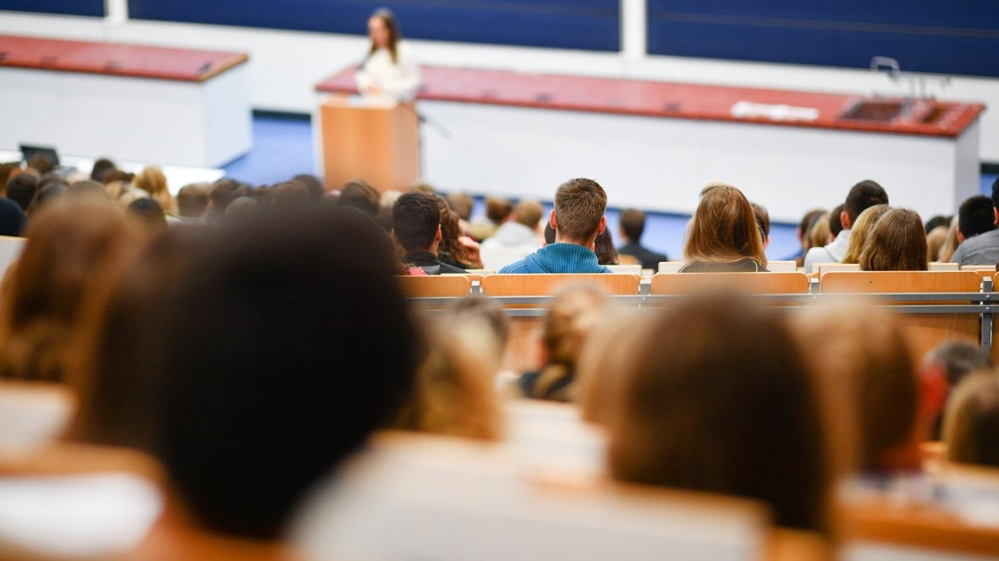 Zahlreiche Studierende sitzen in einem Hörsaal, vorn steht eine Frau am Pult und spricht. 