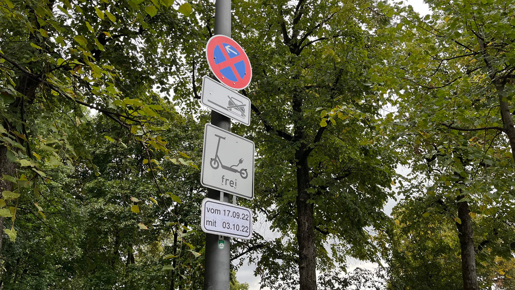 An einem Pfosten sind mehrere Straßenschilder angebracht. Ein Schild mit einem E-Scooter darauf macht deutlich, dass hier E-Scooter geparkt werden dürfen. Darunter ist ein Schild, auf dem steht "vom 17.09.22 mit 03.10.22". 