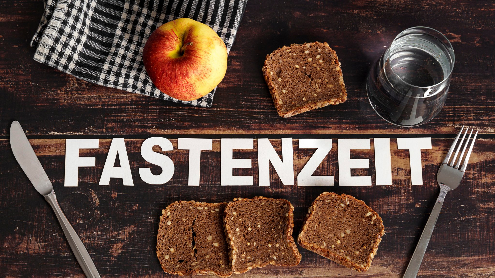 Brot, Äpfel, ein Glas Wasser auf einem Tisch und der Schriftzug "Fastenzeit"