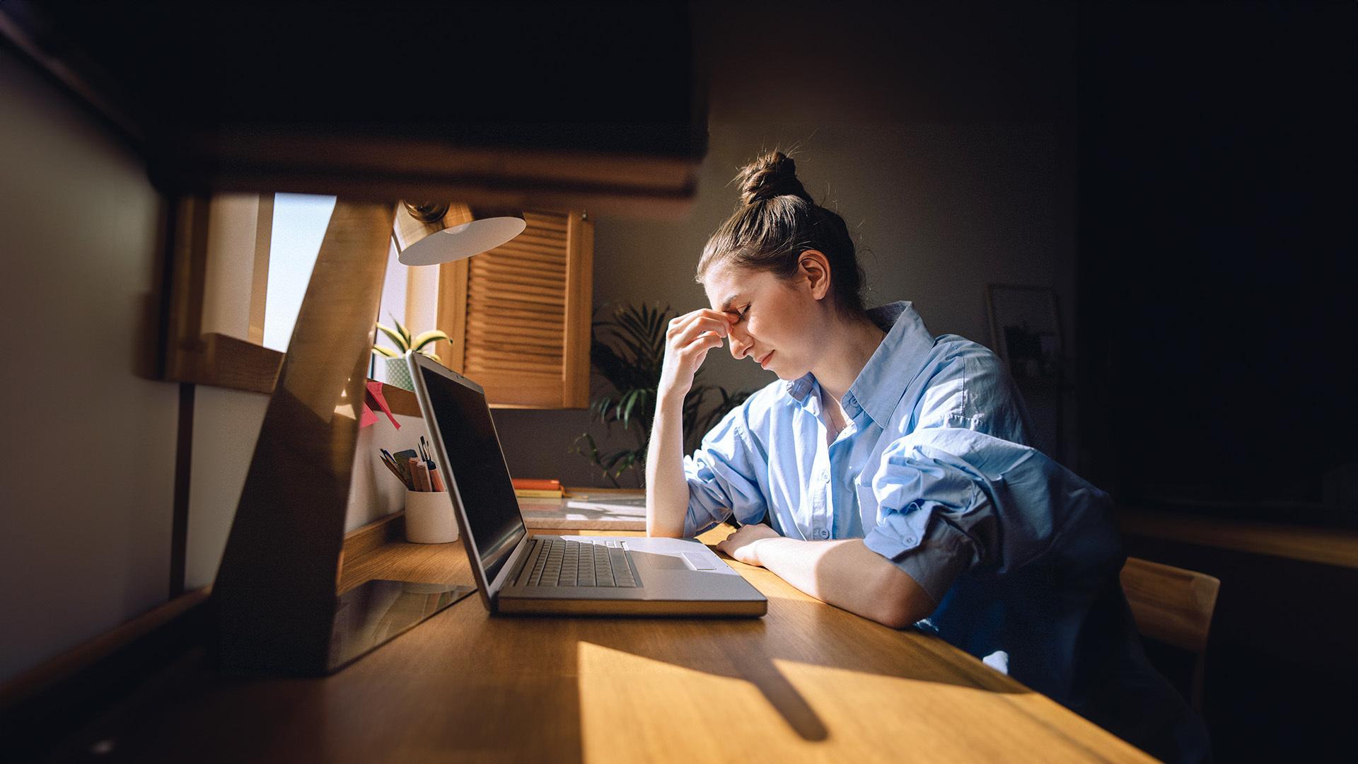 Eine Frau mit Erschöpfungssymptomen sitzt vor einem Laptop an einem Schreibtisch.